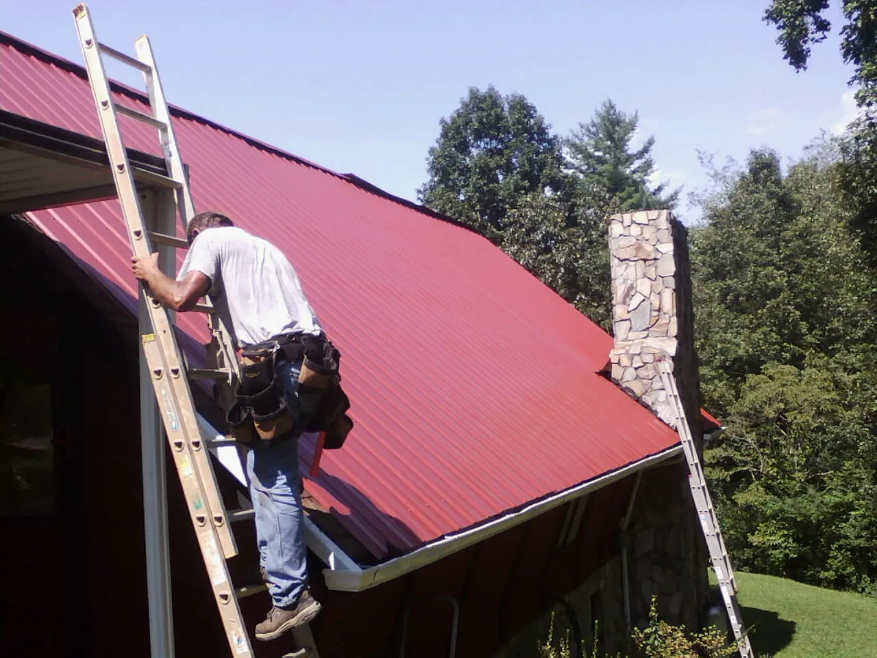 Roofer on a ladder beside a red metal roof and stone chimney — Soffit & Fascia Repair by Roofing Brothers in Lynchburg